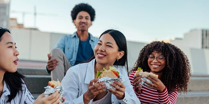 A group of 4 college students are sitting and smiling. 3 of them are eating sandwiches.