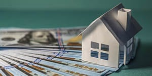 Image of small paper house, sitting on stacks of hundred dollar bills