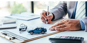 Photo of a person signing a contract with a small toy car, calculator, and car keys on their desk