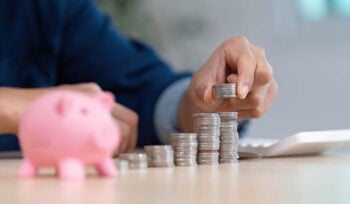 close-up of hands putting coins into a pink piggy bank, with a calculator in the backgroun