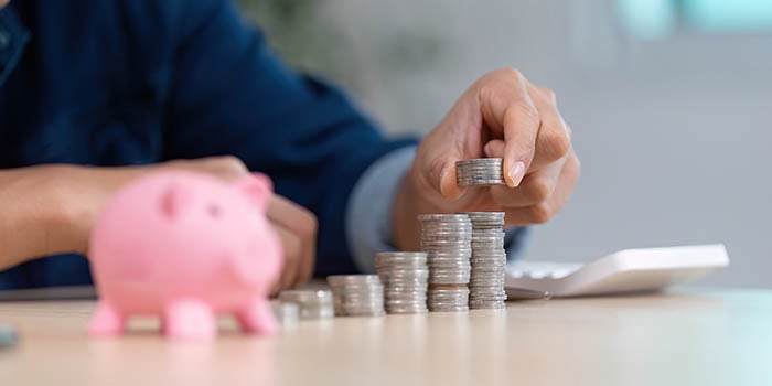 close-up of hands putting coins into a pink piggy bank, with a calculator in the backgroun