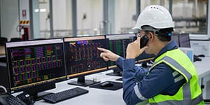 A man in a safety helmet and vest is pointing at one of three computer screens in front of him while also speaking on the phone.