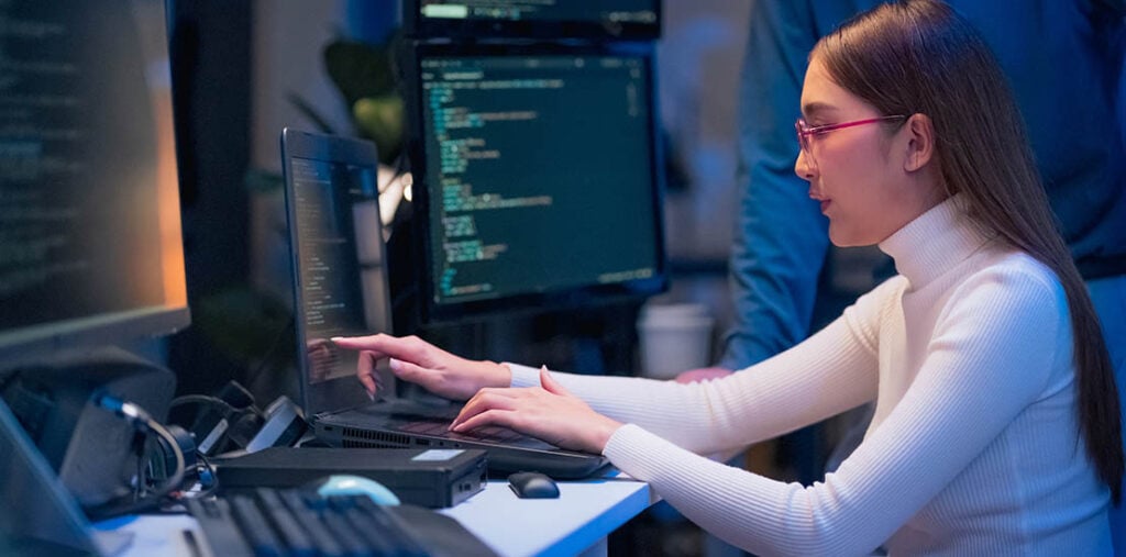A woman sits at a desk with 3 computer monitors, she is pointing at one monitor while typing on her keyboard.