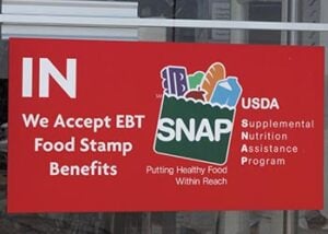 Sign on door at store says "IN We Accept EBT Food Stamp Benefits, with an illustration of a grocery bag with the acronym SNAP on it and with food in it. Other text says "Putting Health Food Within Reach" and USDA Supplemental Nutrition Assistance Program