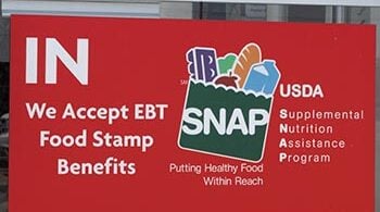 Sign on door at store says "IN We Accept EBT Food Stamp Benefits, with an illustration of a grocery bag with the acronym SNAP on it and with food in it. Other text says "Putting Health Food Within Reach" and USDA Supplemental Nutrition Assistance Program