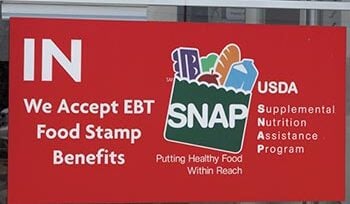Sign on door at store says "IN We Accept EBT Food Stamp Benefits, with an illustration of a grocery bag with the acronym SNAP on it and with food in it. Other text says "Putting Health Food Within Reach" and USDA Supplemental Nutrition Assistance Program