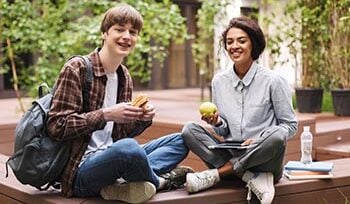 Two college-aged students are sitting and smiling, one is holding a sandwich, the other is holding an apple.