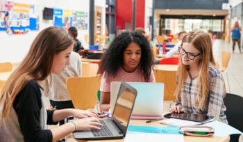 Three college-aged women sit at a desk in a library, looking at their laptops.