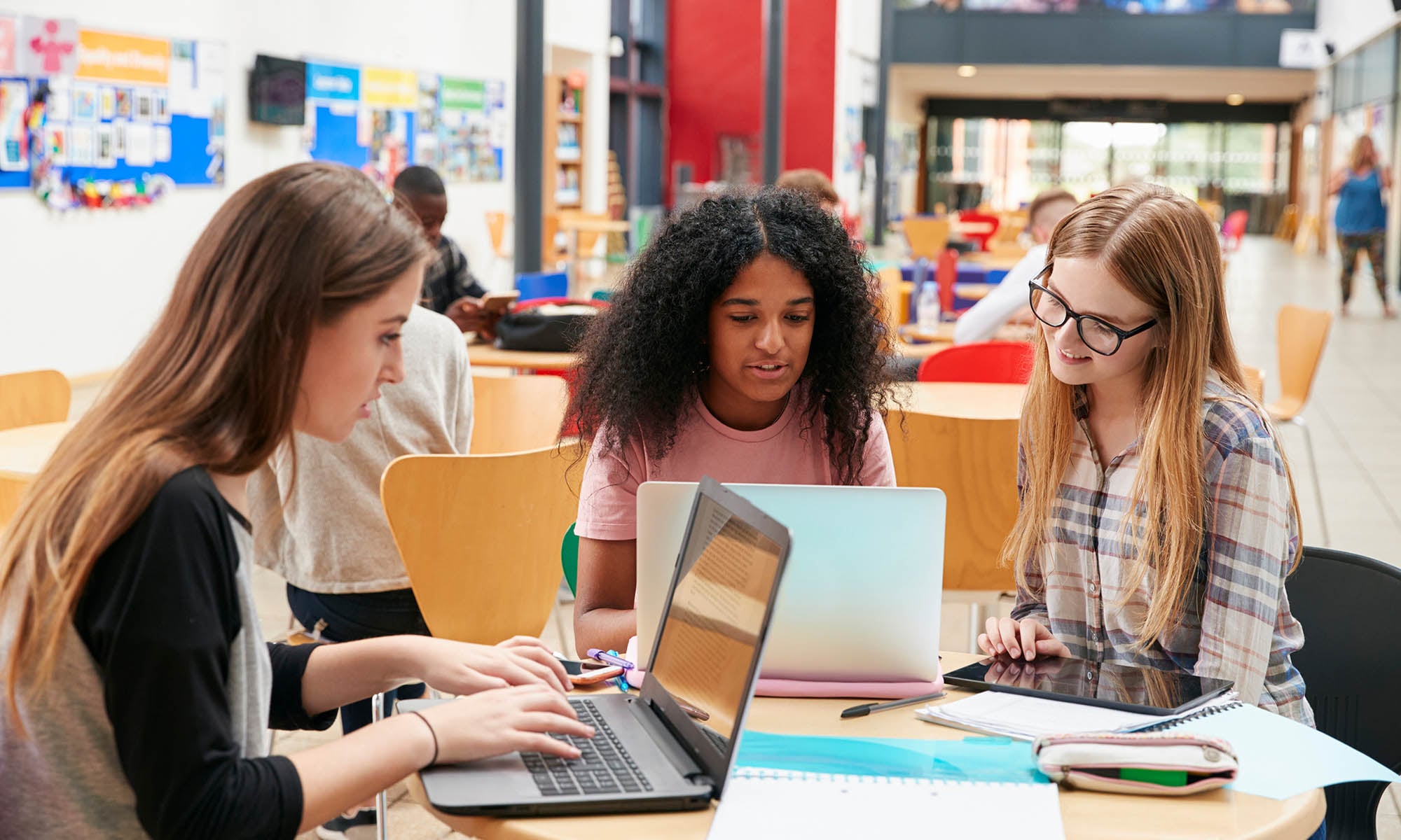 Three college-aged women sit at a desk in a library, looking at their laptops.