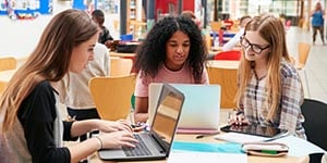 Three college-aged women sit at a desk in a library, looking at their laptops.