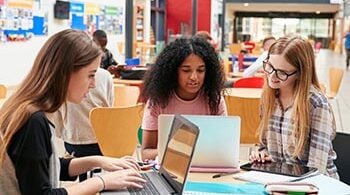 Three college-aged women sit at a desk in a library, looking at their laptops.