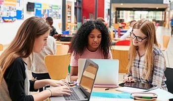 Three college-aged women sit at a desk in a library, looking at their laptops.