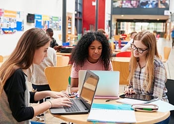 Three college-aged women sit at a desk in a library, looking at their laptops.