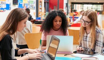 Three college-aged women sit at a desk in a library, looking at their laptops.
