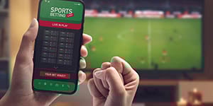 Photograph of person holding a cell phone with a gambling app open with a TV in the background of a sports game.