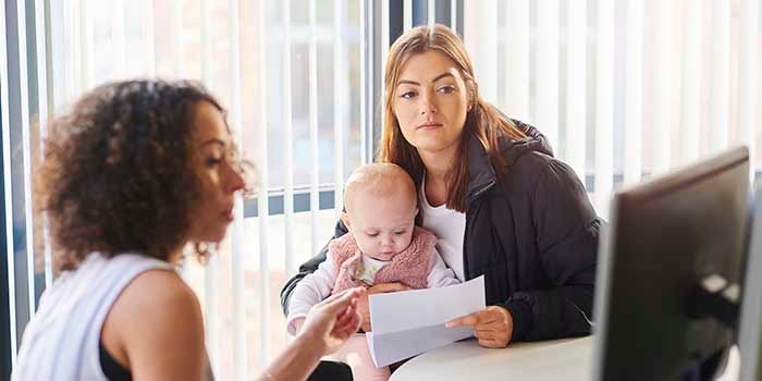 A young mother sits in a chair holding her baby, while speaking with a case manager who is pointing to a computer screen.