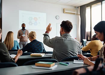 Photo of a college classroom, with several students at desks and professor in font of the room.