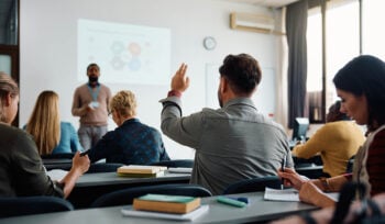 Photo of a college classroom with a student raising his hand to answer a question from the teacher