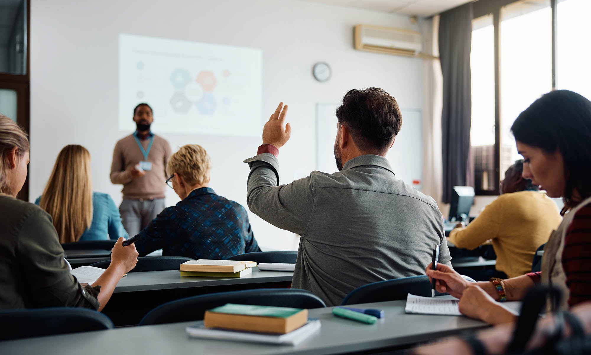 Photo of a college classroom with a student raising his hand to answer a question from the teacher