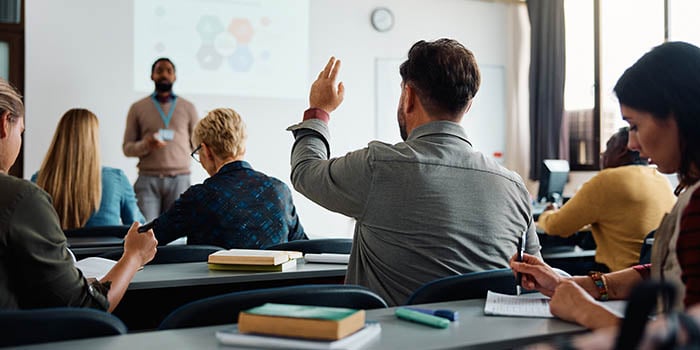 Photo of a college classroom, with several students at desks and professor in font of the room.