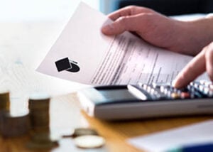 Photo of a man holding a letter related to higher education (with a graduation cap icon on it), while also punching in numbers on a calculator. There are several stacks of change by the calculator.
