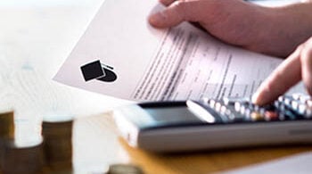 Photo of a man holding a letter related to higher education (with a graduation cap icon on it), while also punching in numbers on a calculator. There are several stacks of change by the calculator.
