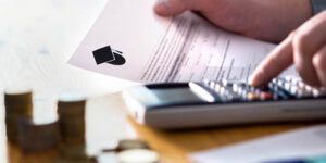 Photo of a man holding a letter related to higher education (with a graduation cap icon on it), while also punching in numbers on a calculator. There are several stacks of change by the calculator.
