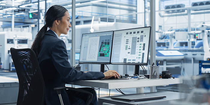 A woman is sitting in an office at a desk with two computers screens - in the background is an industrial/robotic factory.