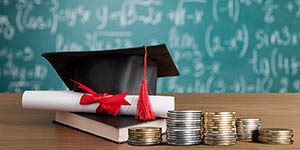 A graduation cap sits on a desk with a diploma, book, and some stacks of change. A blackboard with equations is in the background.