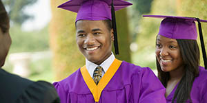 Photo of a college graduate, he's smiling and accepting his diploma. A female college graduate is next to him, also smiling.