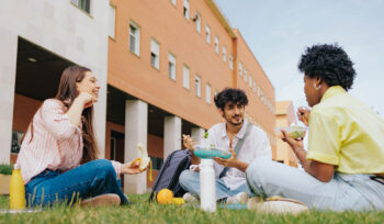 Three college students sit on a lawn outside a building, eating and speaking with each other.