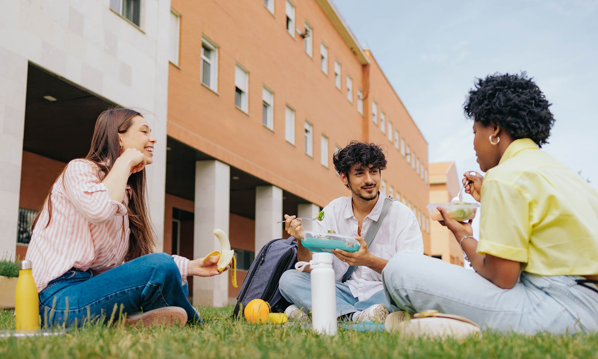Three college students sit on a lawn outside a building, eating and speaking with each other.