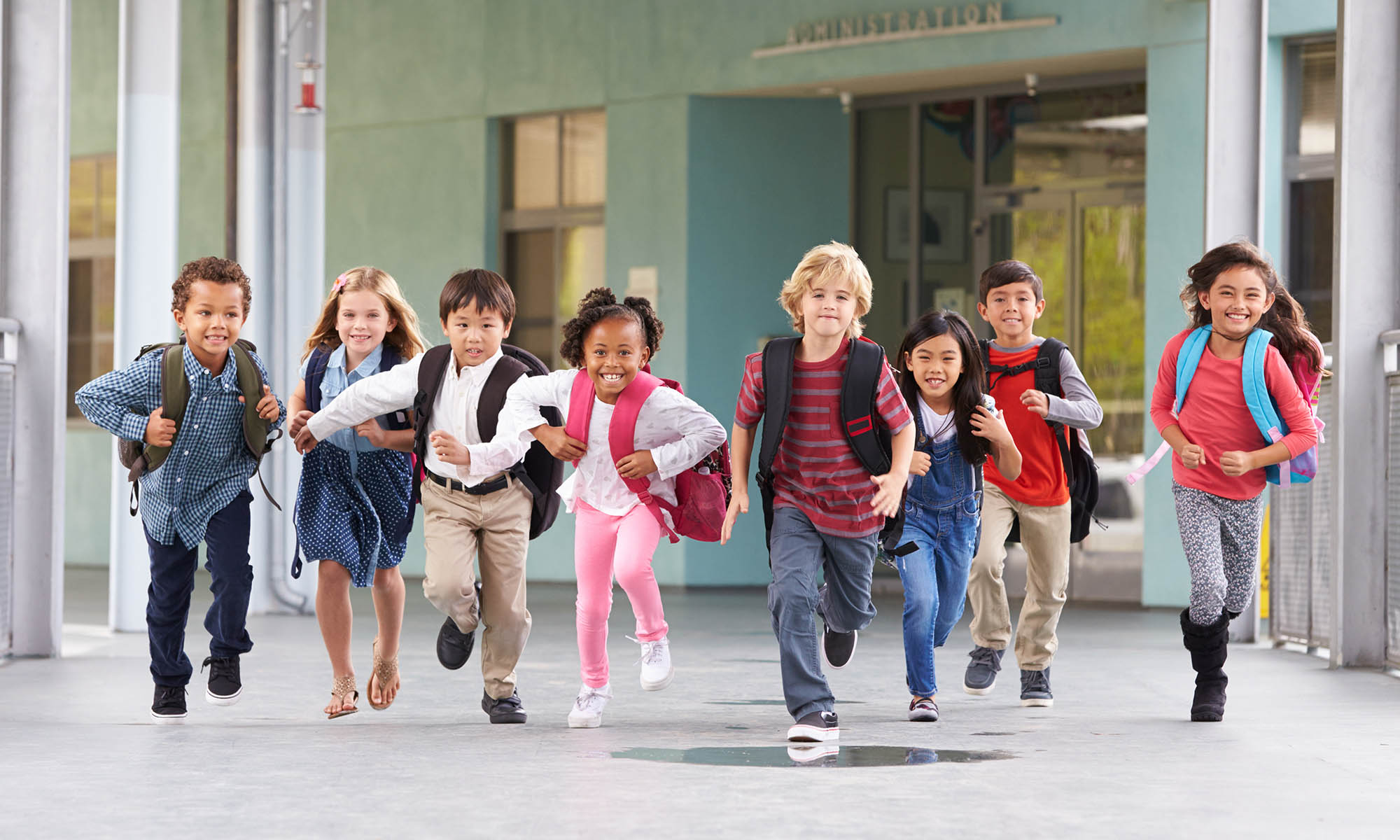 A group of 8 elementary school children are smiling and running in a outdoor school corridor.