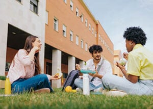 Three college students sit on a lawn outside a building, eating and speaking with each other.