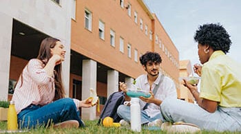 Three college students sit on a lawn outside a building, eating and speaking with each other.