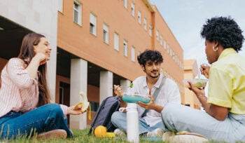 Three college students sit on a lawn outside a building, eating and speaking with each other.