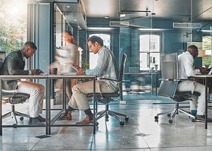 Photograph of an open office space, with three people sitting at desks and a person walking by.
