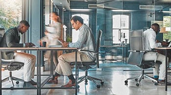 Photograph of an open office space, with three people sitting at desks and a person walking by.