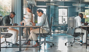 Photograph of an open office space, with three people sitting at desks and a person walking by.