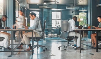 Photograph of an open office space, with three people sitting at desks and a person walking by.