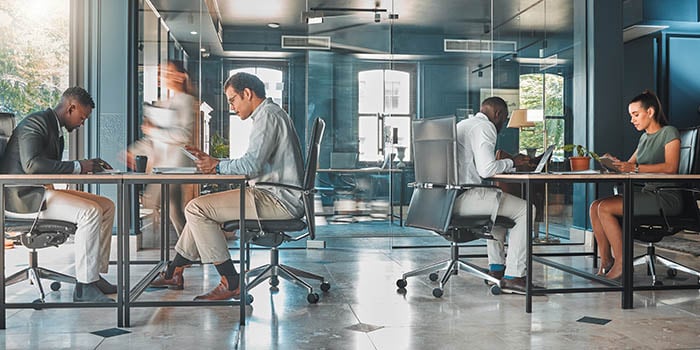 Photograph of an open office space, with three people sitting at desks and a person walking by.