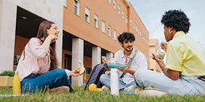 Three college students sit on a lawn outside a building, eating and speaking with each other.