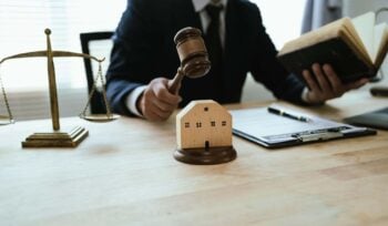 Photo of a judge holding a gavel, with the scales of justice in the background. He's holding a book in his other hand and there is paper and a pen in front of him.