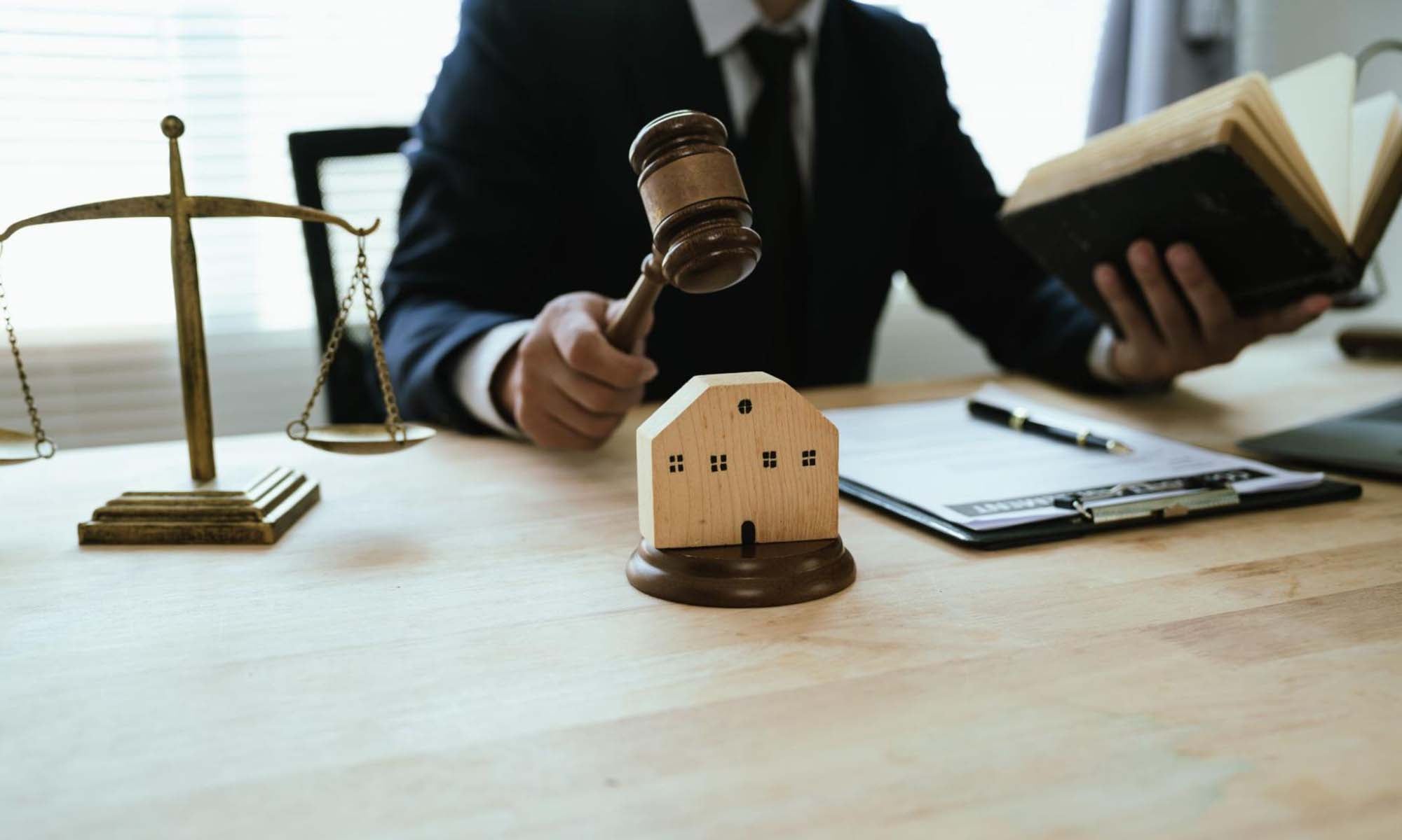 Photo of a judge holding a gavel, with the scales of justice in the background. He's holding a book in his other hand and there is paper and a pen in front of him.