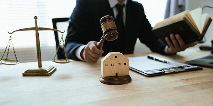 Photo of a judge holding a gavel, with the scales of justice in the background. He's holding a book in his other hand and there is paper and a pen in front of him.