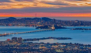 Photograph taken at night, from Oakland, facing towards San Francisco, with the Bay Bridge in the middle and sunset colors in the background