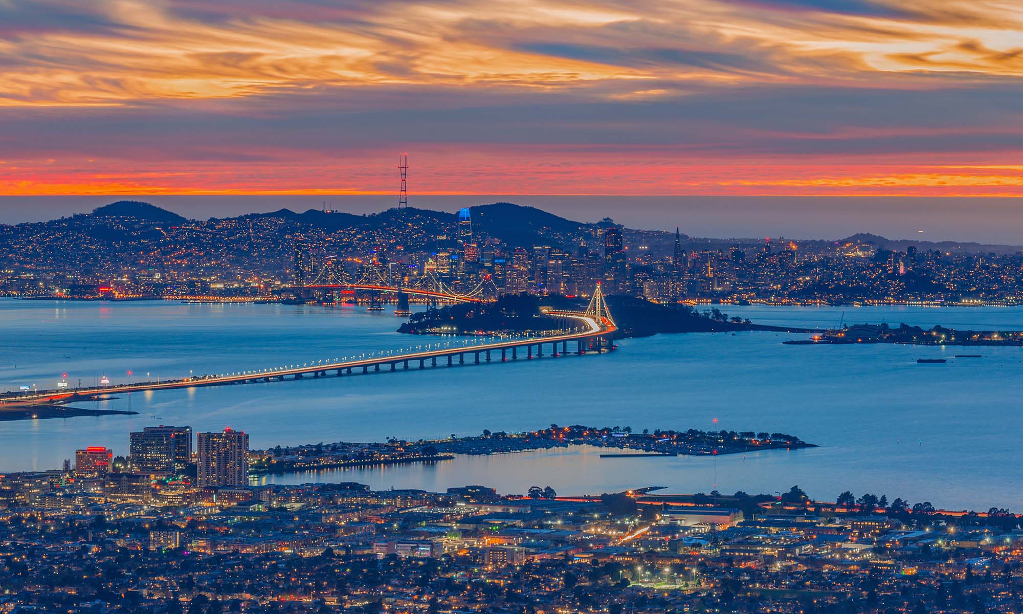 Photograph taken at night, from Oakland, facing towards San Francisco, with the Bay Bridge in the middle and sunset colors in the background