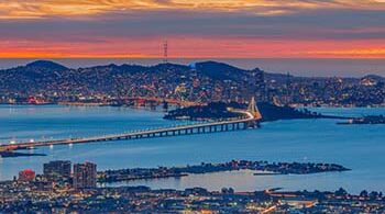 Photograph taken at night, from Oakland, facing towards San Francisco, with the Bay Bridge in the middle and sunset colors in the background