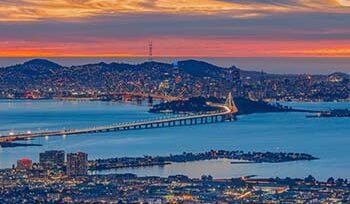 Photograph taken at night, from Oakland, facing towards San Francisco, with the Bay Bridge in the middle and sunset colors in the background