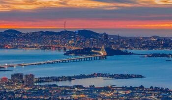 Photograph taken at night, from Oakland, facing towards San Francisco, with the Bay Bridge in the middle and sunset colors in the background